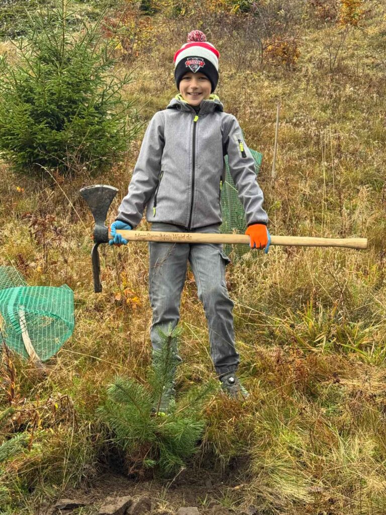 Ein Junge steht stolz mit einer Axt neben einem eingepflanzten Baum.